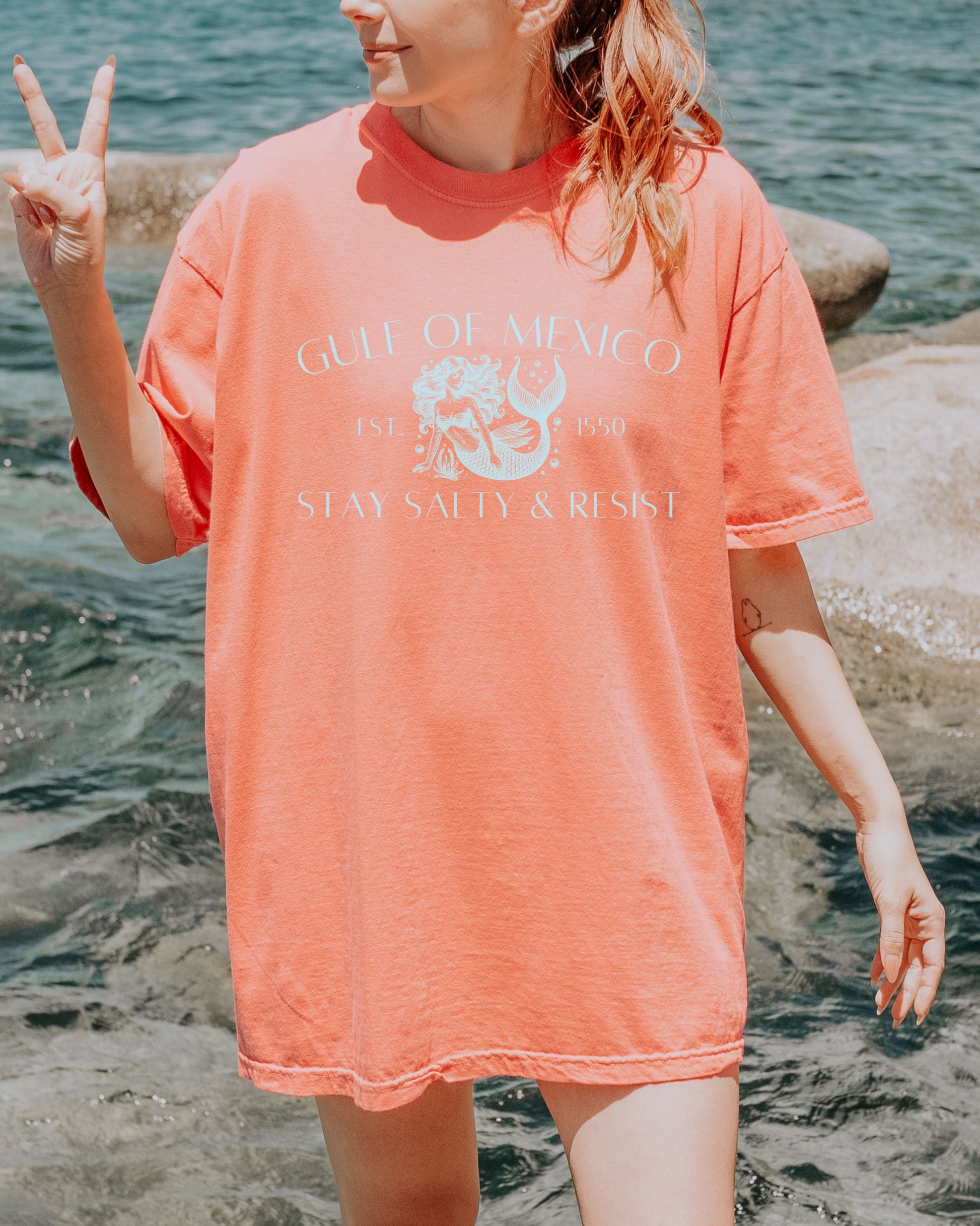 Person wearing a coral t-shirt with text and graphics by the water