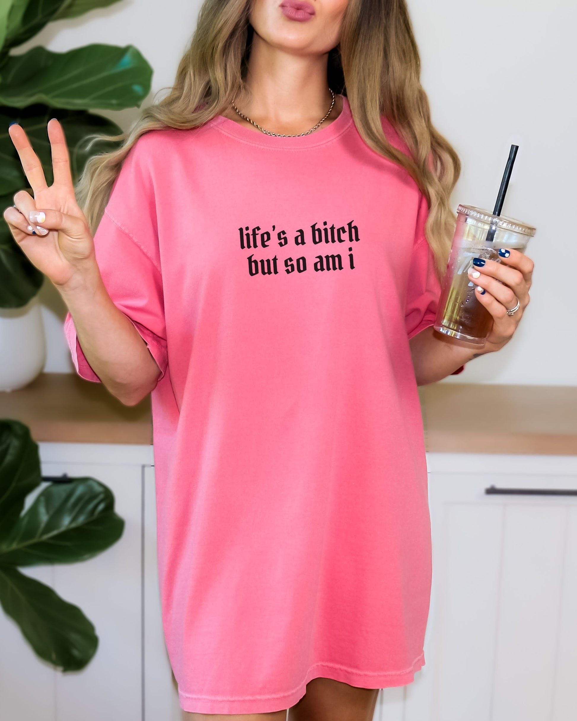 Person wearing a pink shirt with text, making a peace sign and holding a drink.