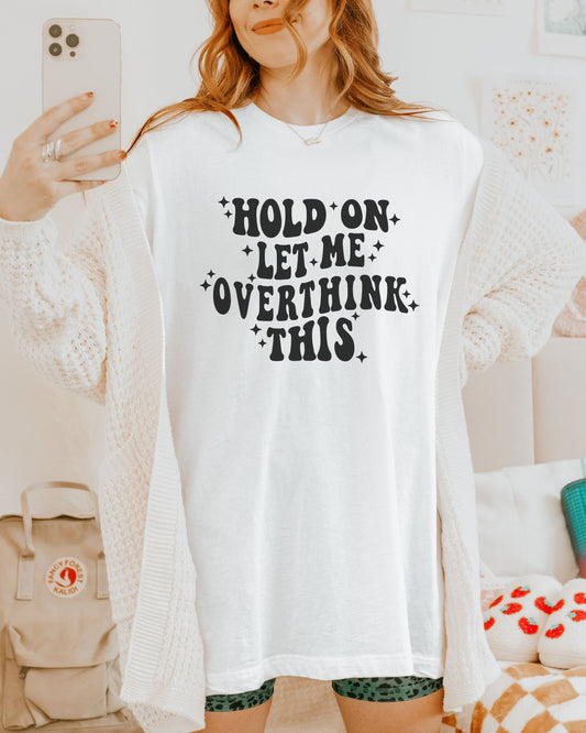 Person wearing a white sweatshirt with text 'Hold on, let me overthink this' in a casual setting.
